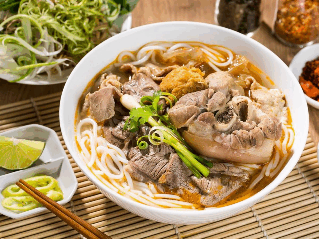 Traditional bowl of Bún Bò Huế - spicy Vietnamese beef noodle soup from Hue, Central Vietnam, served with fresh herbs, chili, and lime.