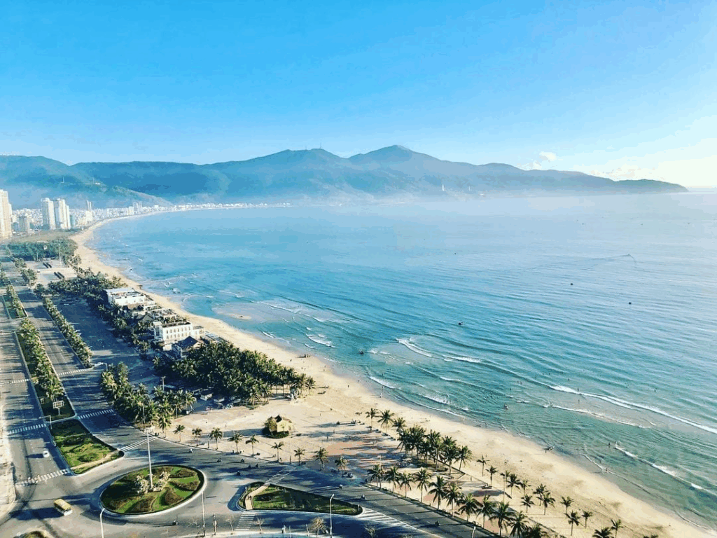 Aerial view of My Khe Beach in Da Nang, Central Vietnam - a long sandy coastline lined with palm trees, clear blue water, and the Son Tra mountains in the background.