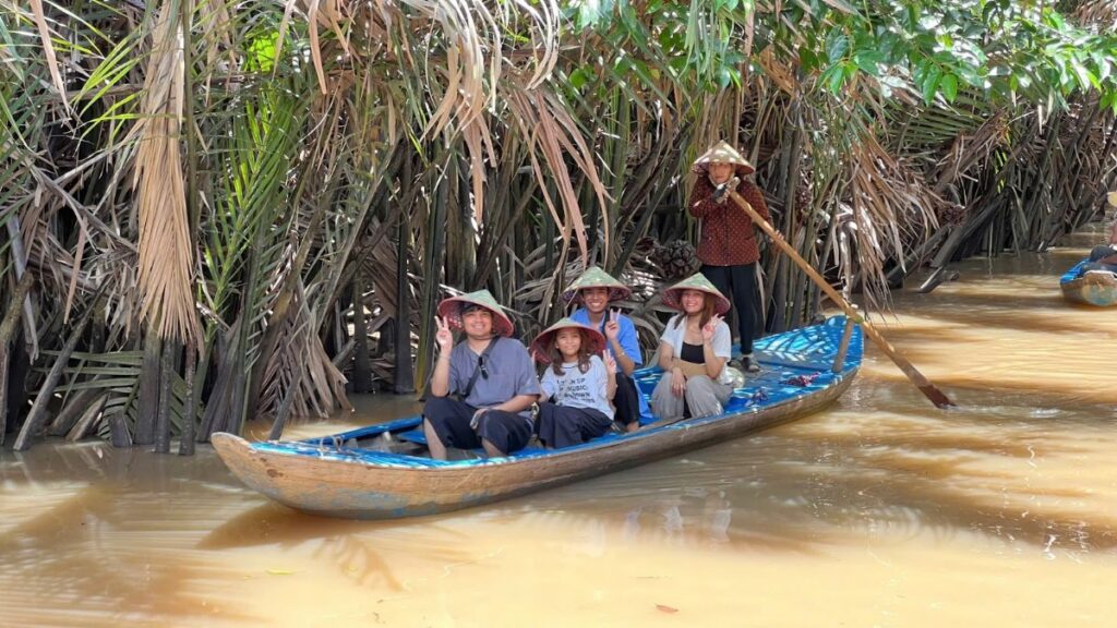 Guests in conical hats riding a wooden sampan through a palm-lined canal on a Mekong Delta day trip from Saigon