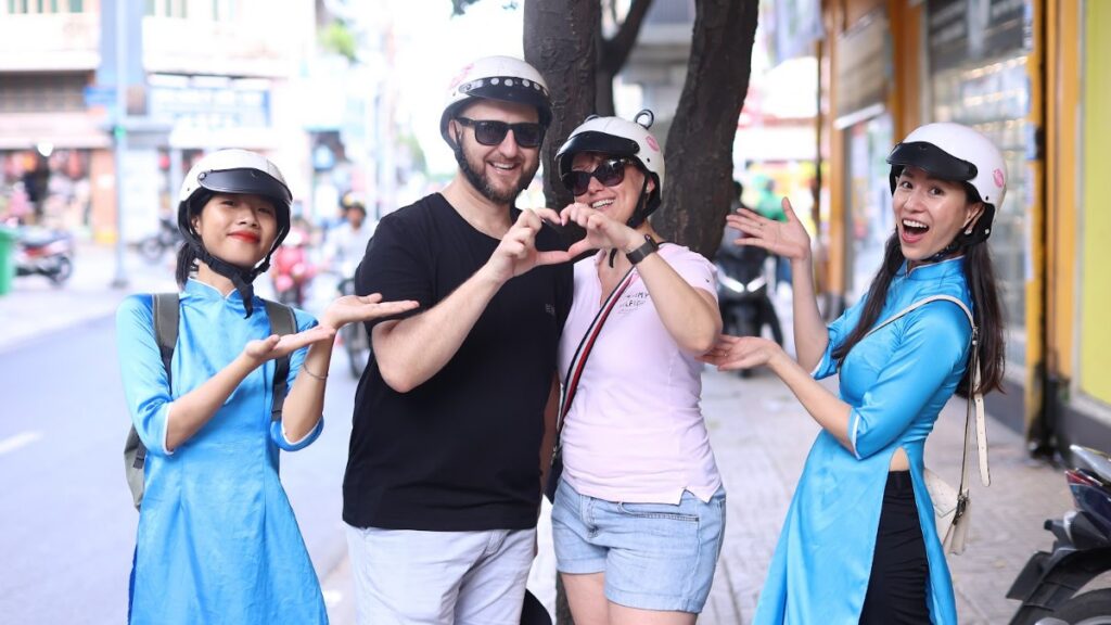 Travellers in shorts and t-shirts wearing helmets with KissTour guides in ao dai on a Saigon street, showing what to wear in Vietnam